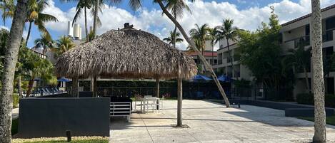 A covered outdoor area with a thatched roof, palm trees, and a building in the background.
