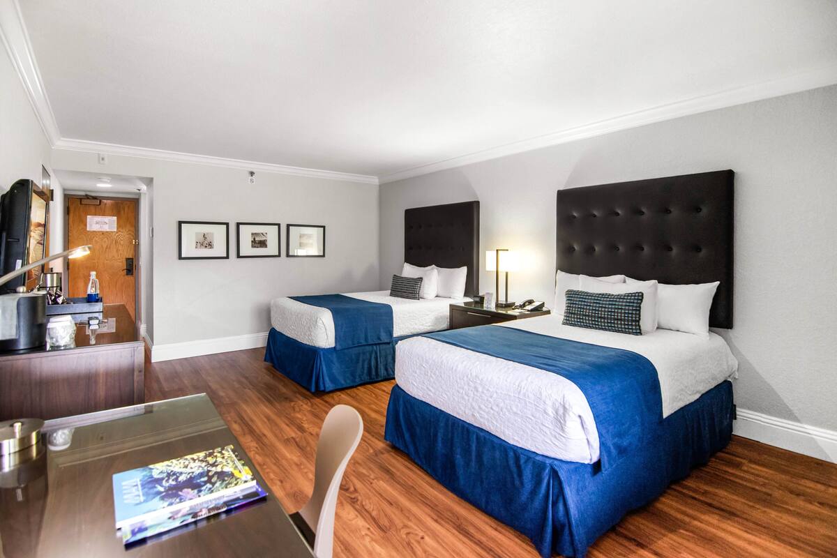 A hotel room with two beds, a desk, and a chair. There are framed pictures on the wall and a lamp on the nightstand.