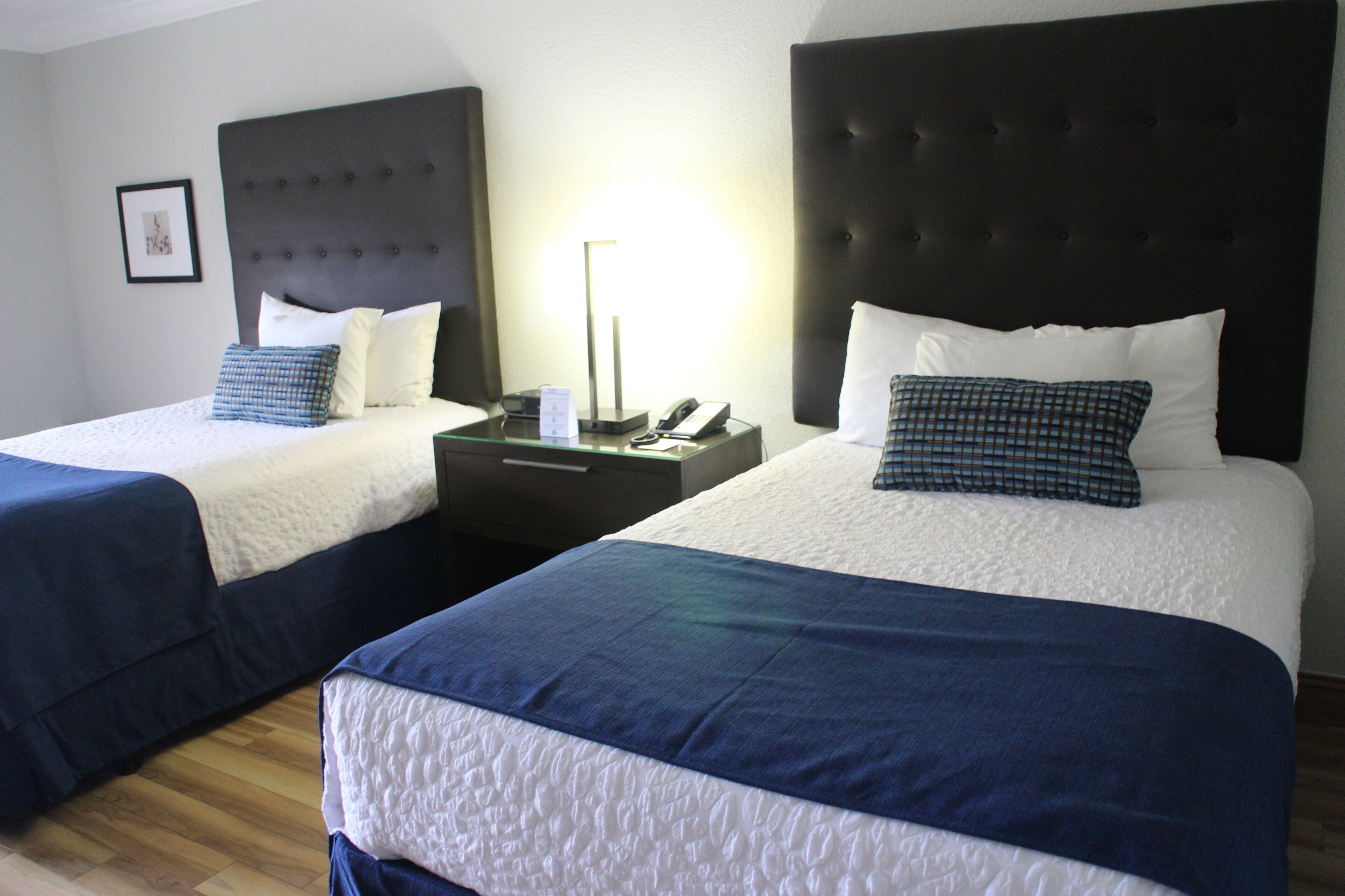 A hotel room with two beds, each with a dark headboard and white bedding. There is a bedside table with a lamp and a phone between the beds.