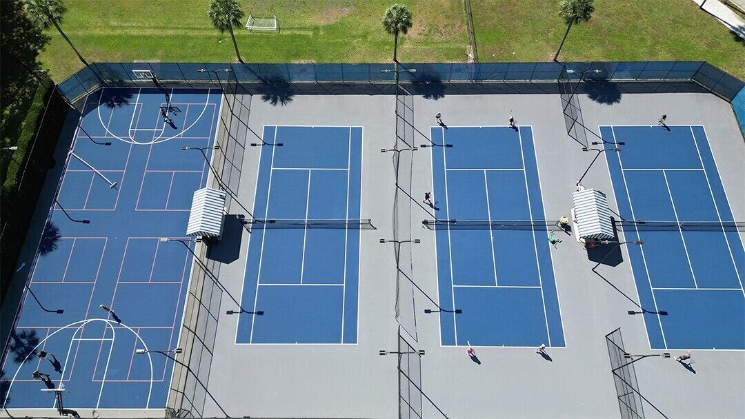 Eine Vogelperspektive auf einen Tennisplatzkomplex mit mehreren Plätzen, einer überdachten Zuschauertribüne und umliegender Bepflanzung.