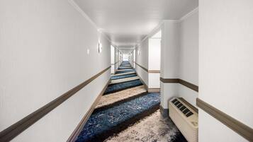 Un pasillo largo y estrecho en un hotel, con paredes blancas, alfombra azul y un equipo de aire acondicionado color beige.