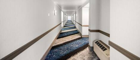 A long, narrow hotel hallway with white walls, a blue carpet, and a beige air conditioning unit.