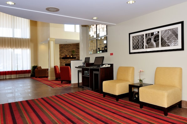 Um saguão de hotel com duas poltronas, um balcão de recepção, um tapete listrado vermelho e uma grande obra de arte emoldurada na parede.