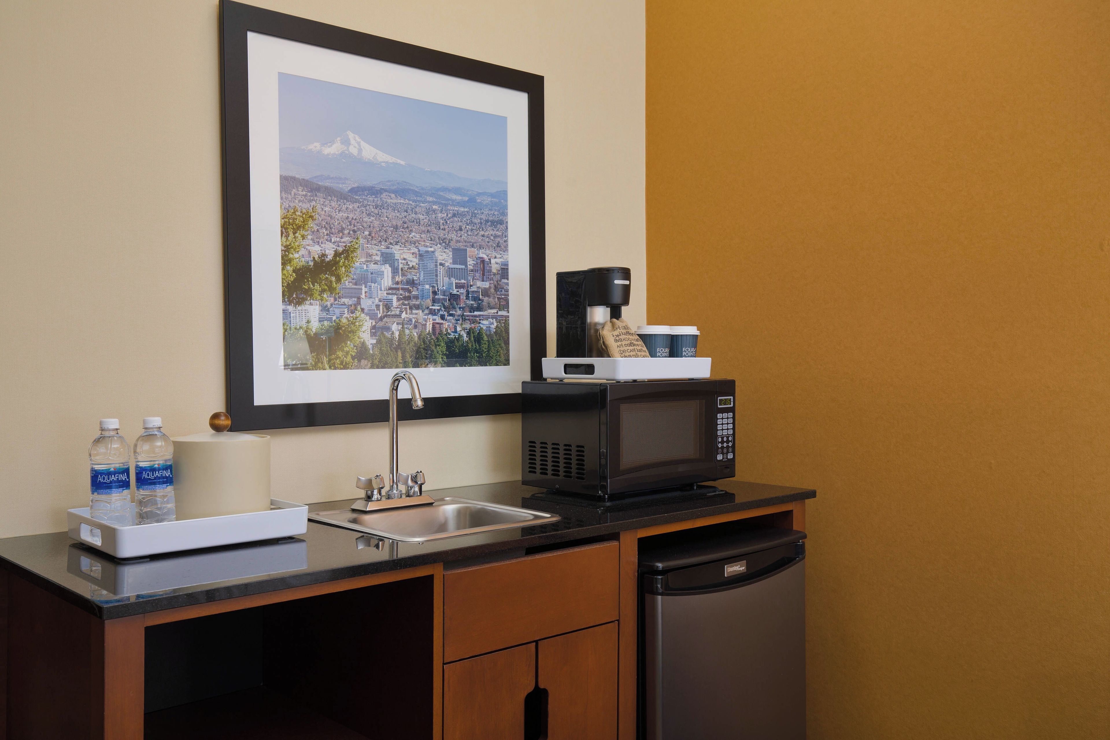 A compact kitchenette with a coffee maker, microwave, sink, and a framed cityscape photo on the wall.