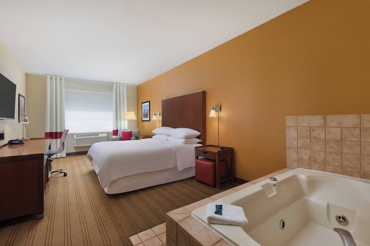 A hotel room with a bed, a desk, a chair, a desk lamp, a bedside table, a lamp, a window with curtains, a wall-mounted TV, and a bathtub.