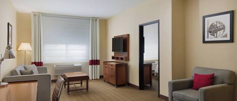 Habitación de hotel con sofá, una silla, escritorio, televisor y un amplio ventanal con cortinas.