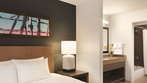 Et hotelværelse med en seng, et natbord med lampe, samt et badeværelse med vask, spejl og brusebad.