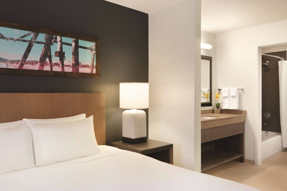 Ein Hotelzimmer mit Bett, Nachttisch mit Lampe, Bad mit Waschbecken, Spiegel und Dusche.