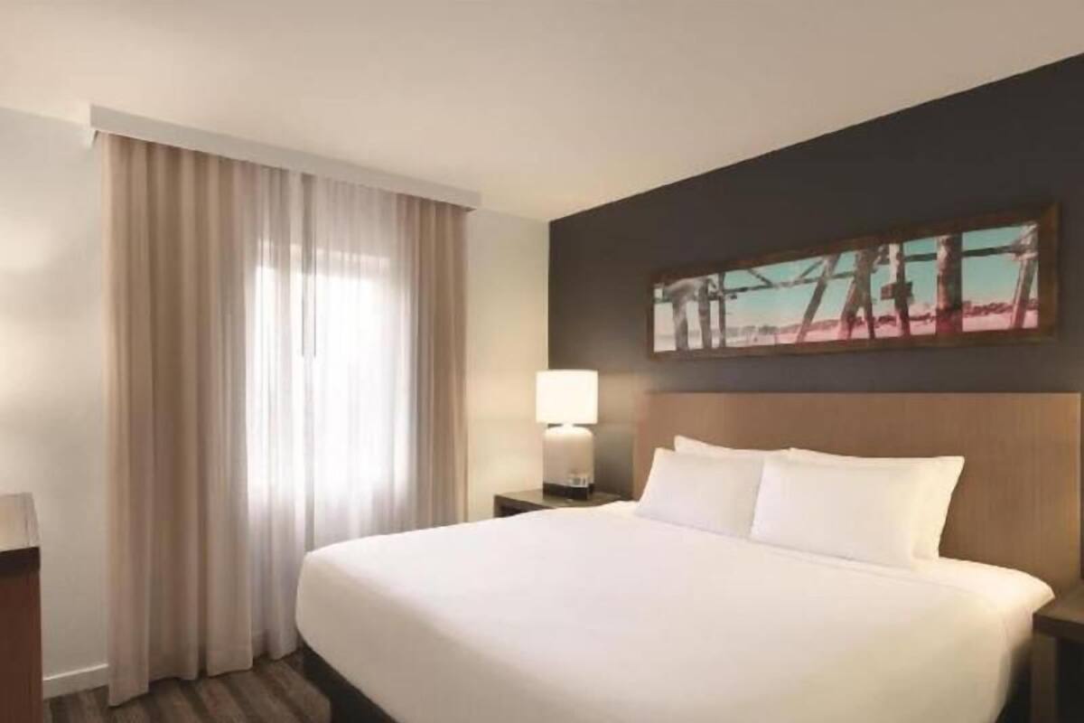 Ein Hotelzimmer mit einem großen Bett, einem Nachttisch mit Lampe, einem Fenster mit Vorhängen und einem Bild an der Wand.