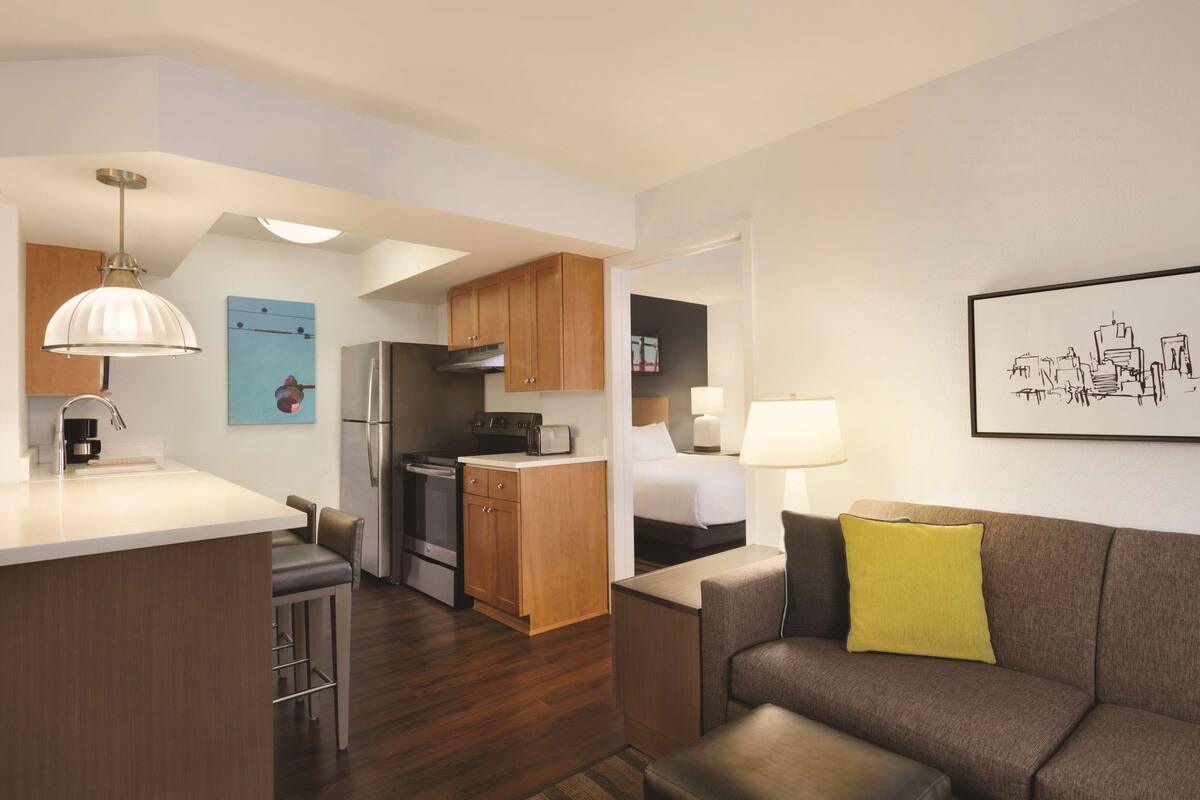 Ein modernes Hotelzimmer mit einer Küchenzeile, einem Sofa mit gelbem Kissen, einem Bett und einem Stadtbild an der Wand.