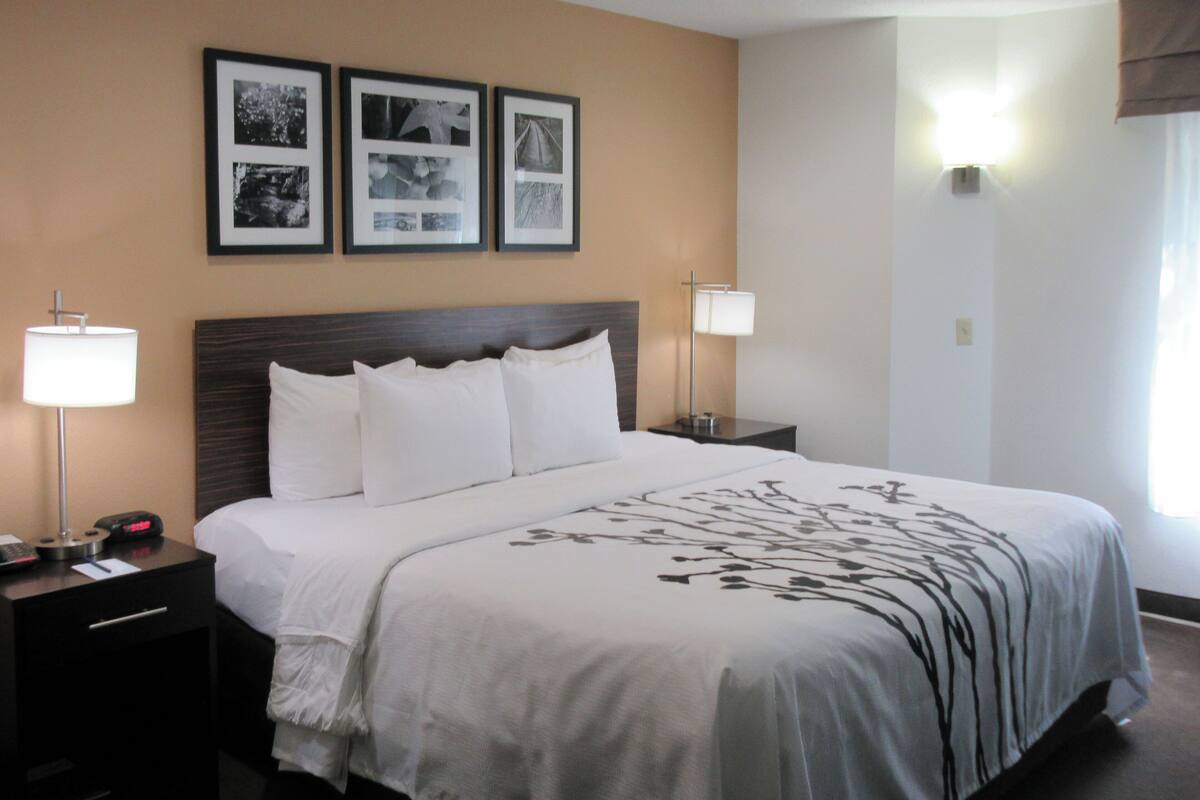 A hotel room with a large bed, two bedside tables, a nightstand, and a wall-mounted lamp. There are three framed pictures on the wall above the bed.