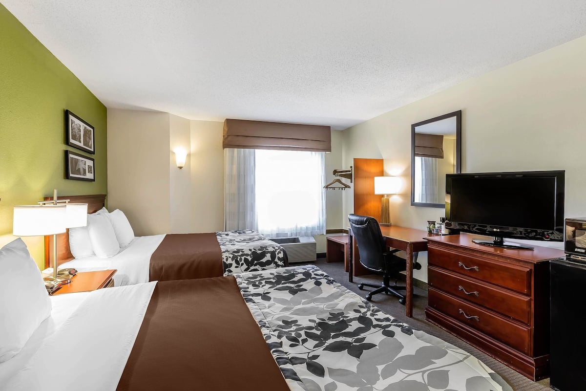 Quarto de hotel com duas camas, uma escrivaninha com televisão, cômoda e espelho.