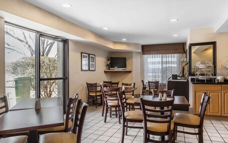A hotel dining area with multiple tables and chairs, a television mounted on a wall, and a kitchen area visible in the background.