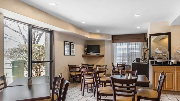 Une salle à manger d’hôtel avec plusieurs tables et chaises, un téléviseur fixé au mur et une cuisine visible au fond.