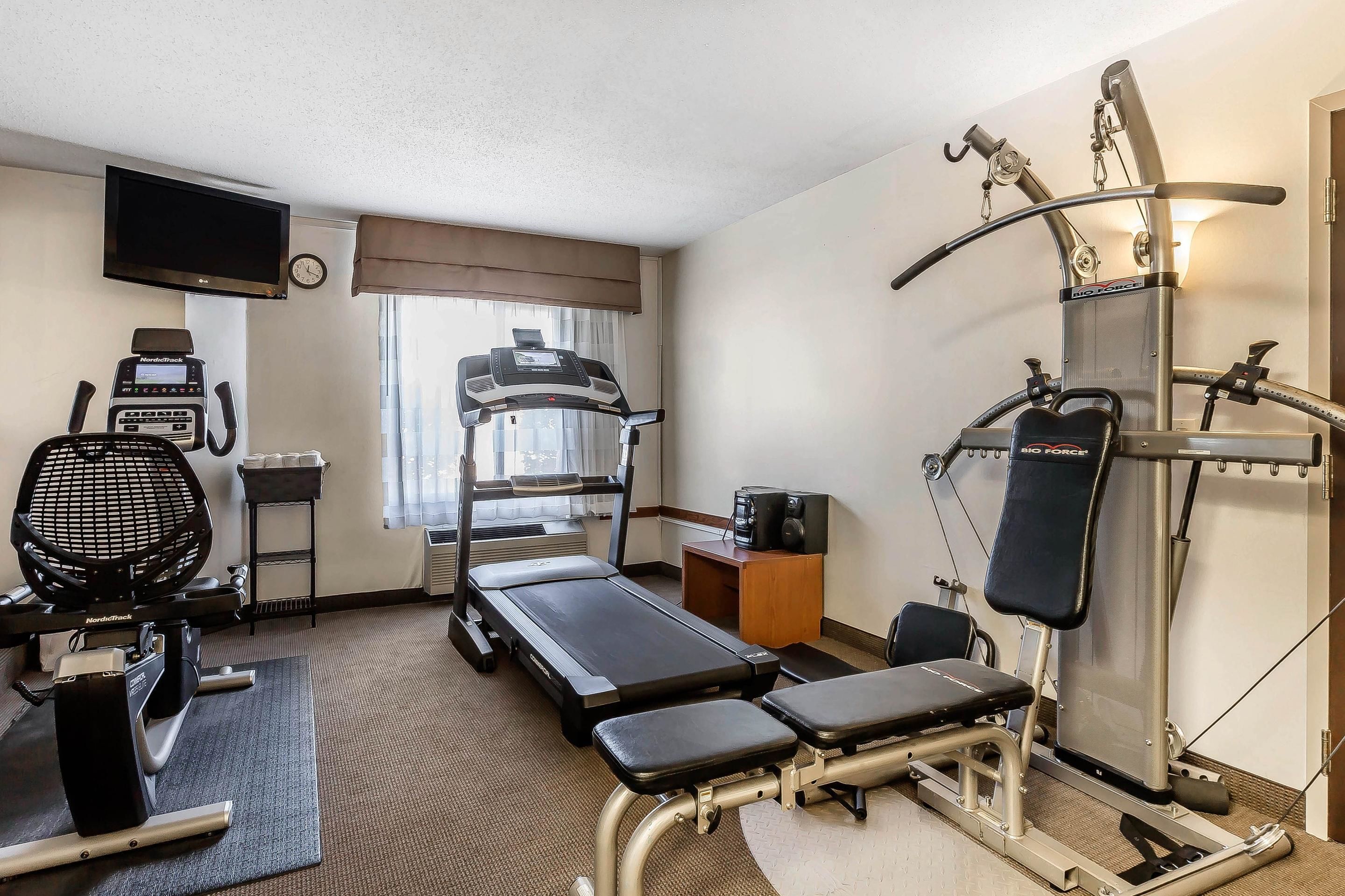 A hotel room with a treadmill, stationary bike, rowing machine, and a TV mounted on the wall.