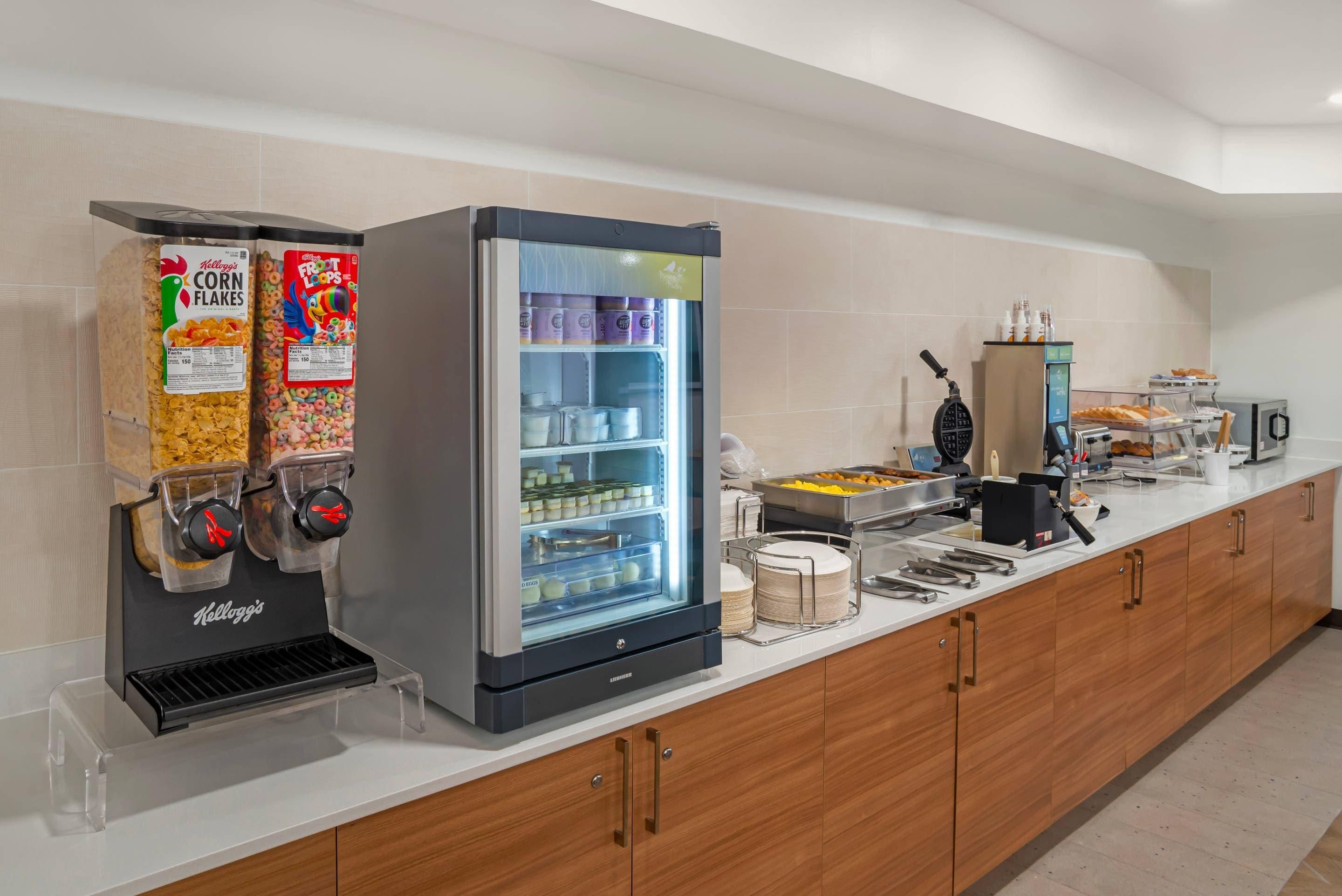 A self-service breakfast area with cereal dispensers, a refrigerated section, and a coffee machine.