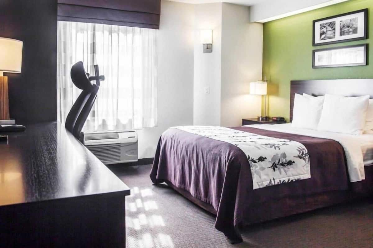 A hotel room with a bed, a desk, a chair, a lamp, and a window with curtains.