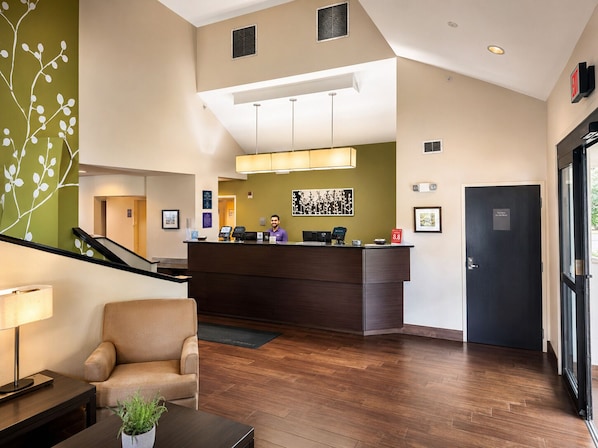 A hotel lobby with a reception desk, a sofa, a coffee table, a plant, and a staircase.