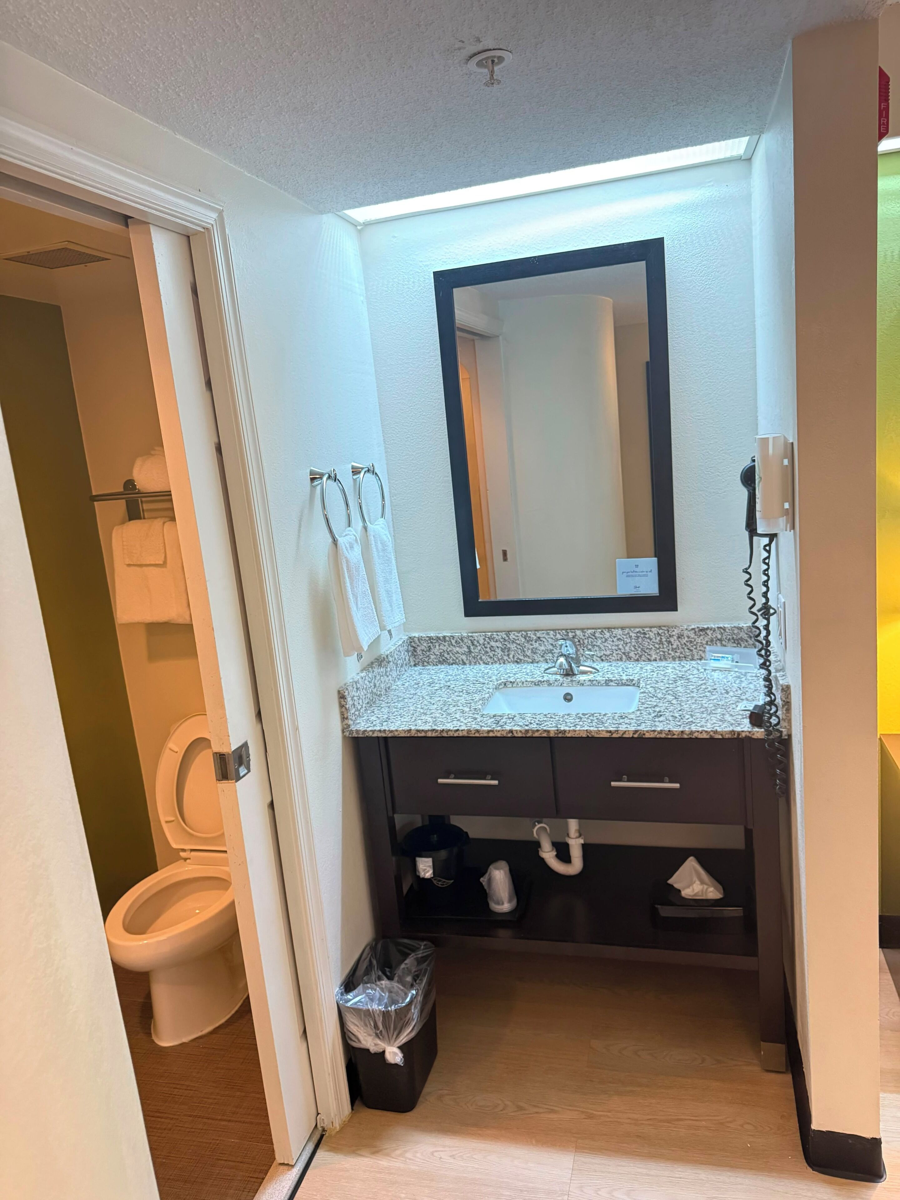 Une salle de bain équipée de toilettes, d’un lavabo, d’un miroir, d’un porte-serviettes et d’une poubelle.