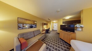 A compact hotel room with a sofa, a small TV, a kitchenette, and a patterned floor. - Residences at International Place (Tewksbury)