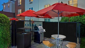 An outdoor patio with a grill, wicker furniture, and red umbrellas. - Residences at International Place (Tewksbury)