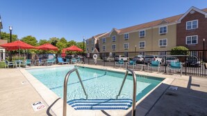 A swimming pool with lounge chairs and umbrellas, surrounded by a fence and a building with multiple windows. - Residences at International Place (Tewksbury)
