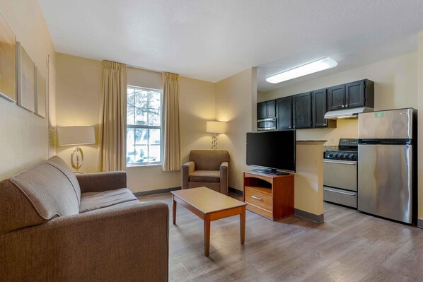 A compact living space with a sofa, a television, a dining table, a kitchen area with cabinets and appliances, and a window with curtains.
