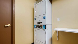 Un appareil combiné lave-linge et sèche-linge, de type « superposé », est installé dans un coin de la pièce, près d’une porte et d’une petite étagère.