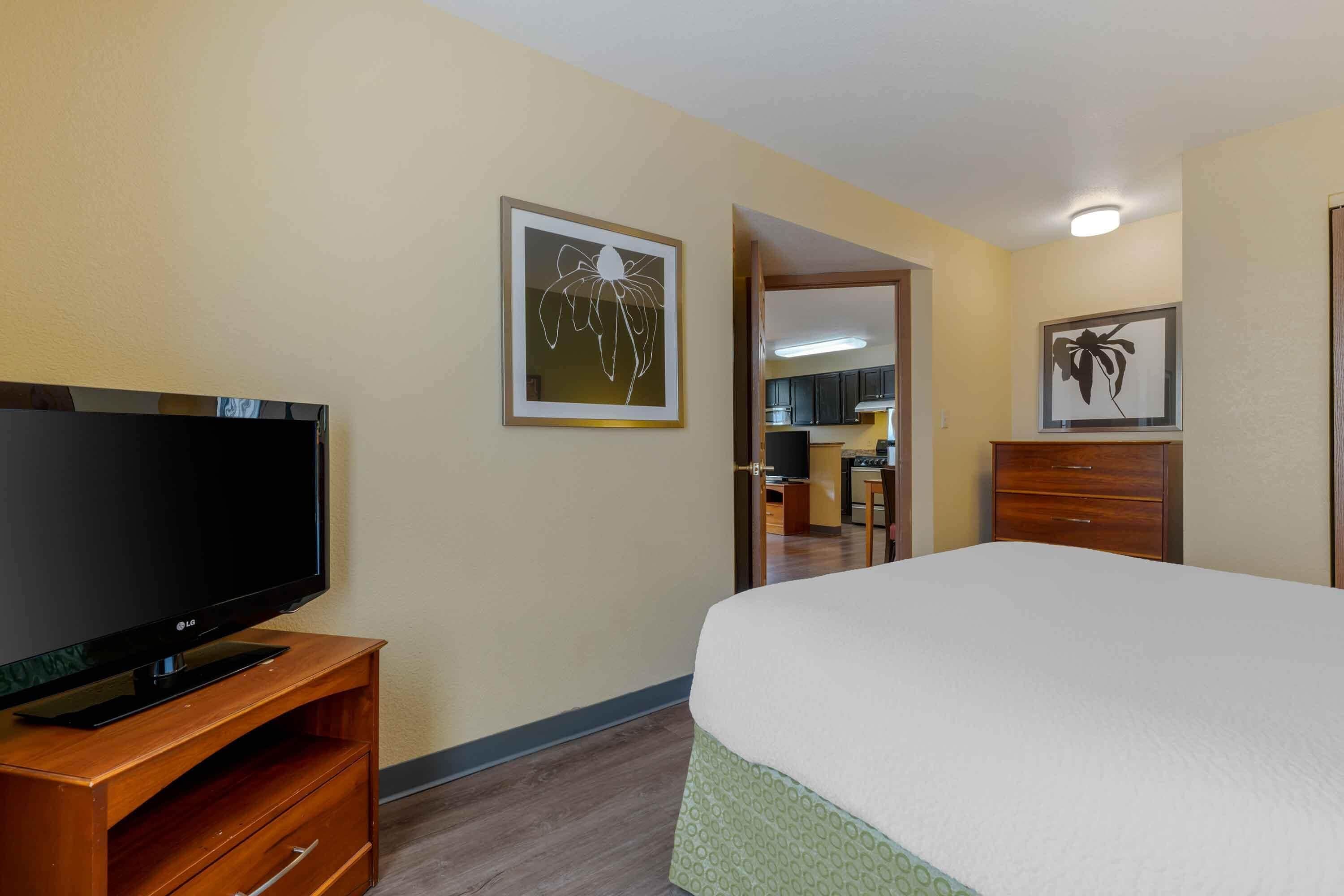 Habitación de hotel con cama, televisor sobre un mueble de madera, puerta que da a un baño y un cuadro en la pared.