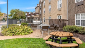 A patio area with a grill, dining table, and chairs, surrounded by a wooden fence and greenery.