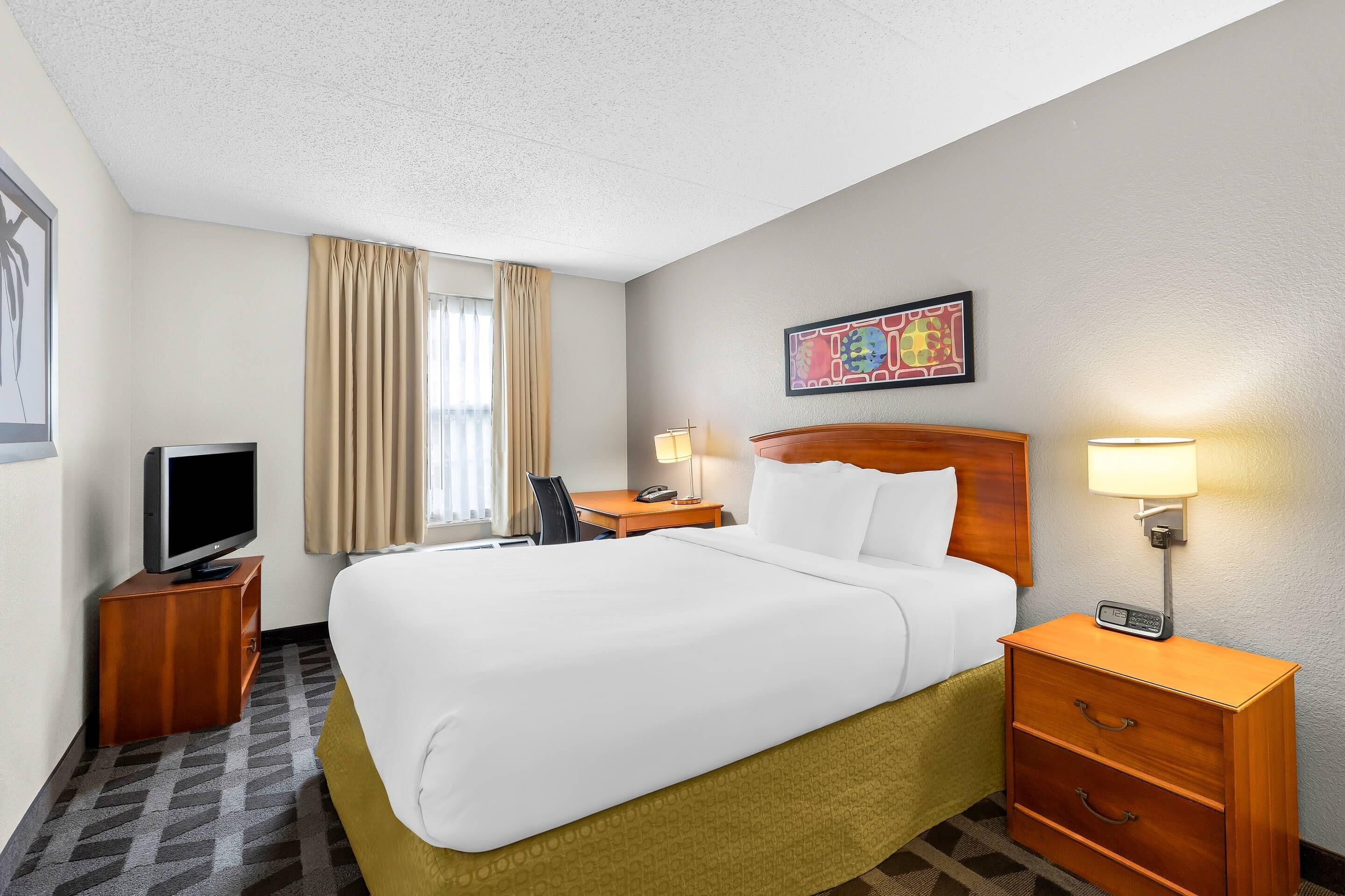 A hotel room with a large bed, a desk with a chair, a television, and a nightstand with a lamp.