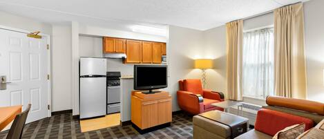 A compact hotel room with a kitchenette, a sofa, a dining table, and a TV.