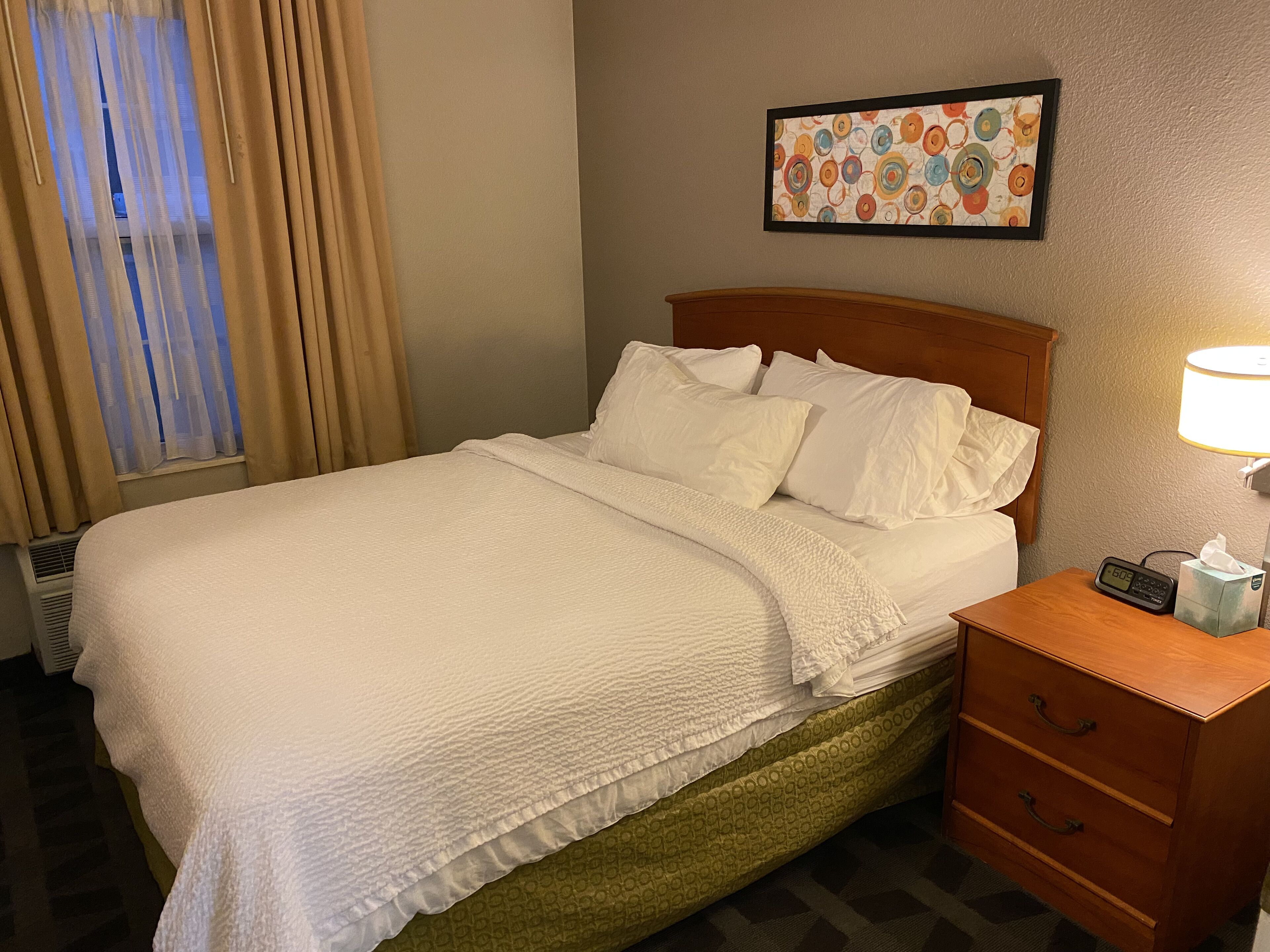 A hotel room with a bed, a bedside table with a lamp, a clock, and a framed artwork on the wall.
