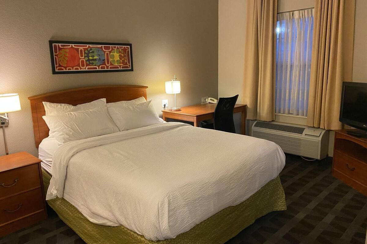 Ett hotellrum med en säng, ett skrivbord, en stol, en tv och ett fönster med gardiner.