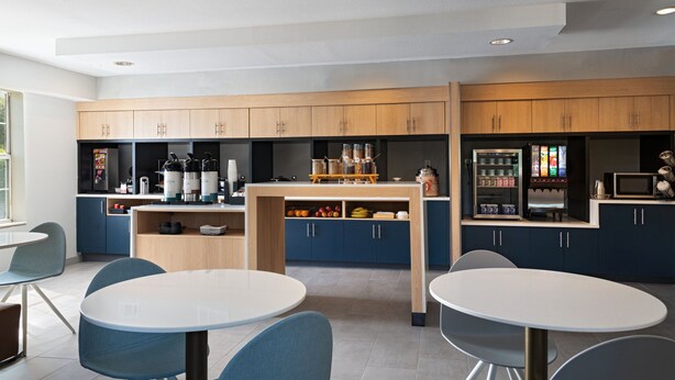 A modern kitchen with wooden cabinets, a central island, and a seating area with round tables and chairs. - TownePlace Suites by Marriott College Station (College Station)