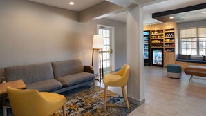 A modern living room with a sofa, chairs, a coffee table, a floor lamp, and a bookshelf with snacks. - TownePlace Suites by Marriott College Station (College Station)