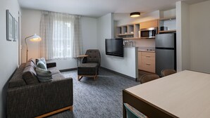 A modern, compact living space with a sofa, armchair, and a dining table. A kitchen area with a microwave, refrigerator, and a flat-screen TV is visible. - TownePlace Suites by Marriott College Station (College Station)