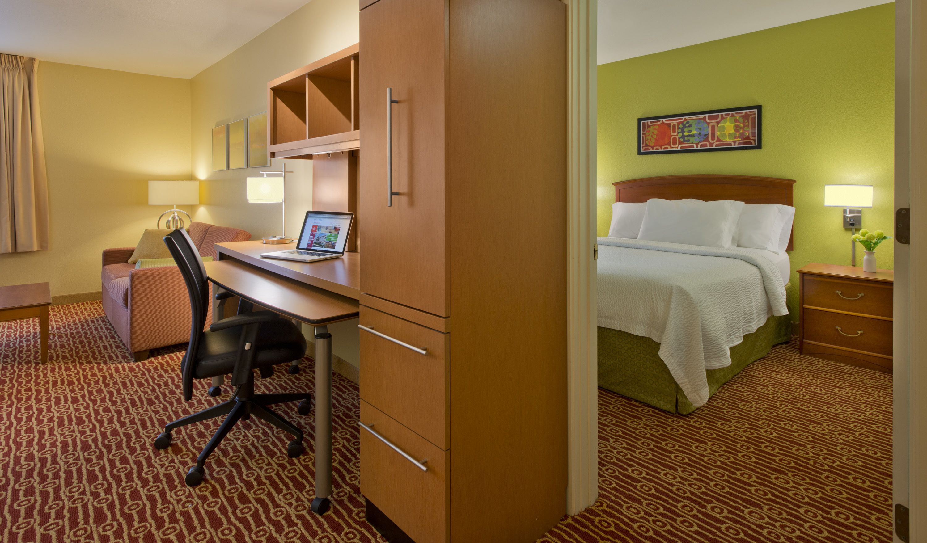 A hotel room with a desk, chair, bed, and a small table with a lamp.