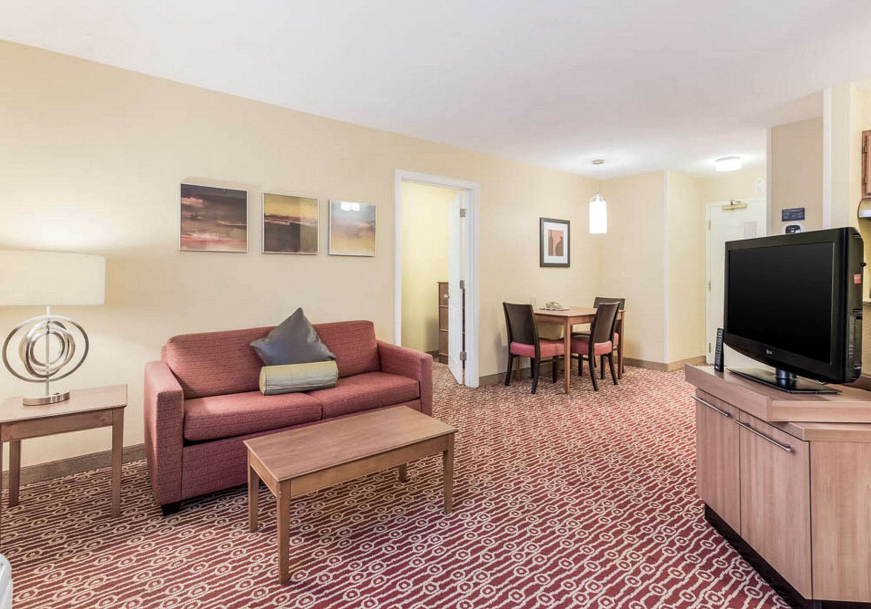 A hotel room with a red patterned carpet, a red sofa with cushions, a wooden coffee table, a TV on a wooden stand, a dining table with chairs, and framed pictures on the wall.