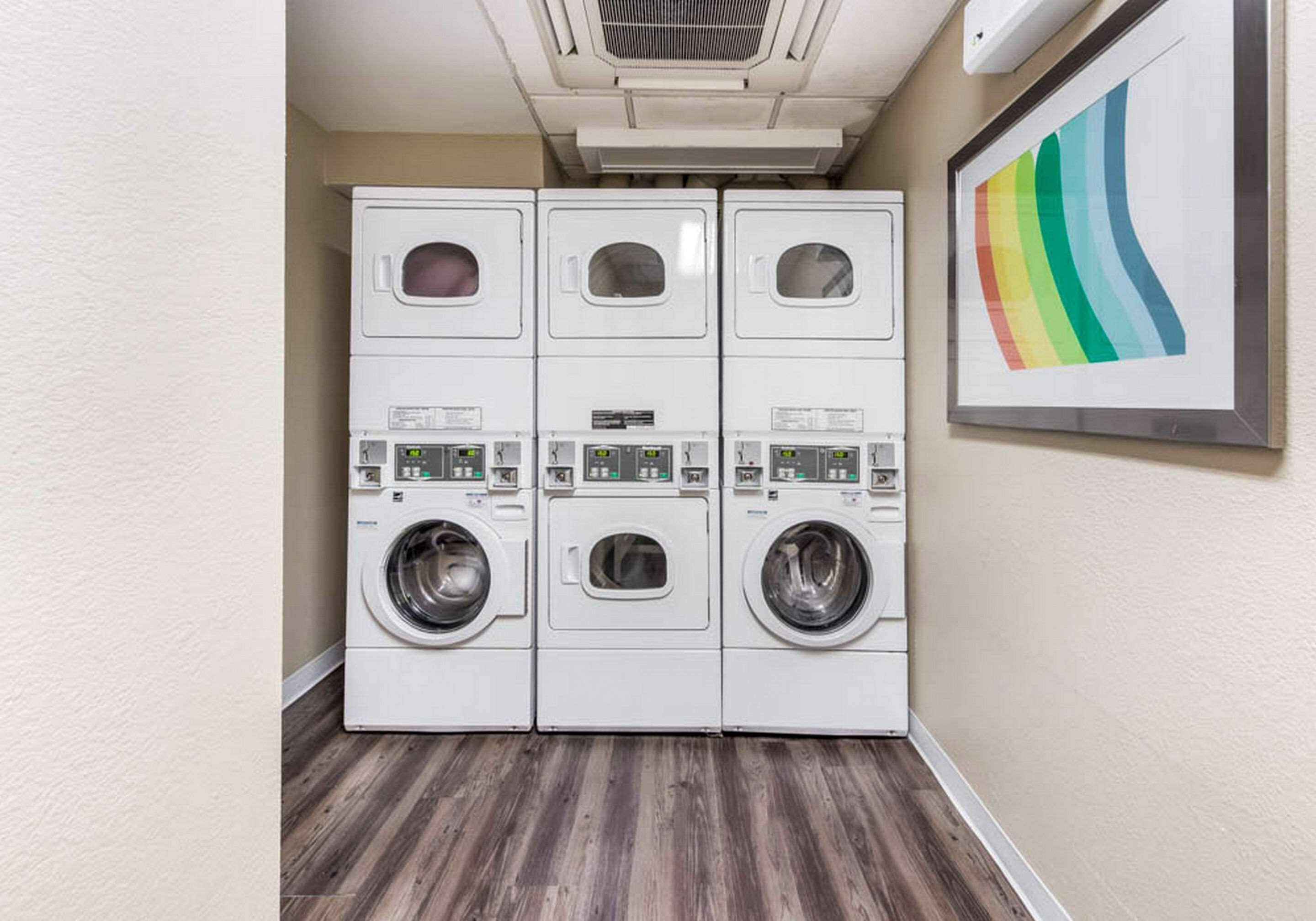 A laundry area with stacked washing machines and dryers, a framed artwork on the wall, and a wooden floor.