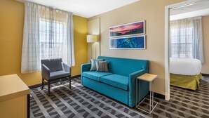 A hotel room with a blue sofa, a small table, a chair, a bed with a green bed skirt, and two paintings on the wall. - MainStay Suites Houston NASA/Clear Lake (Houston)