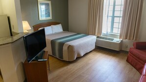 A hotel room with a bed, a TV on a wooden stand, a red chair, a window with curtains, and a wall-mounted picture. - Siegel Select Montgomery (Montgomery)