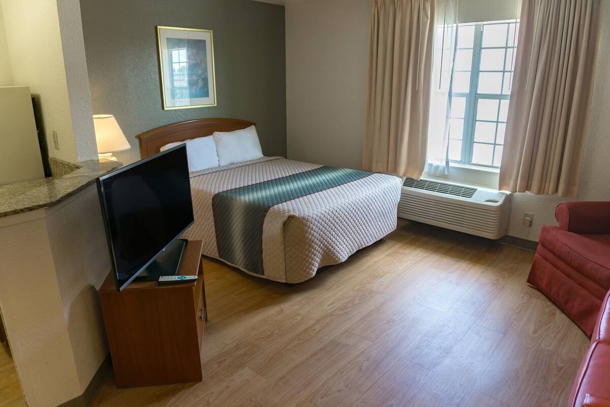 A hotel room with a bed, a TV on a wooden stand, a red chair, a window with curtains, and a wall-mounted picture.