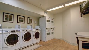 A laundry room with multiple washing machines and dryers, a small table, and framed pictures on the wall. - Siegel Select Montgomery (Montgomery)
