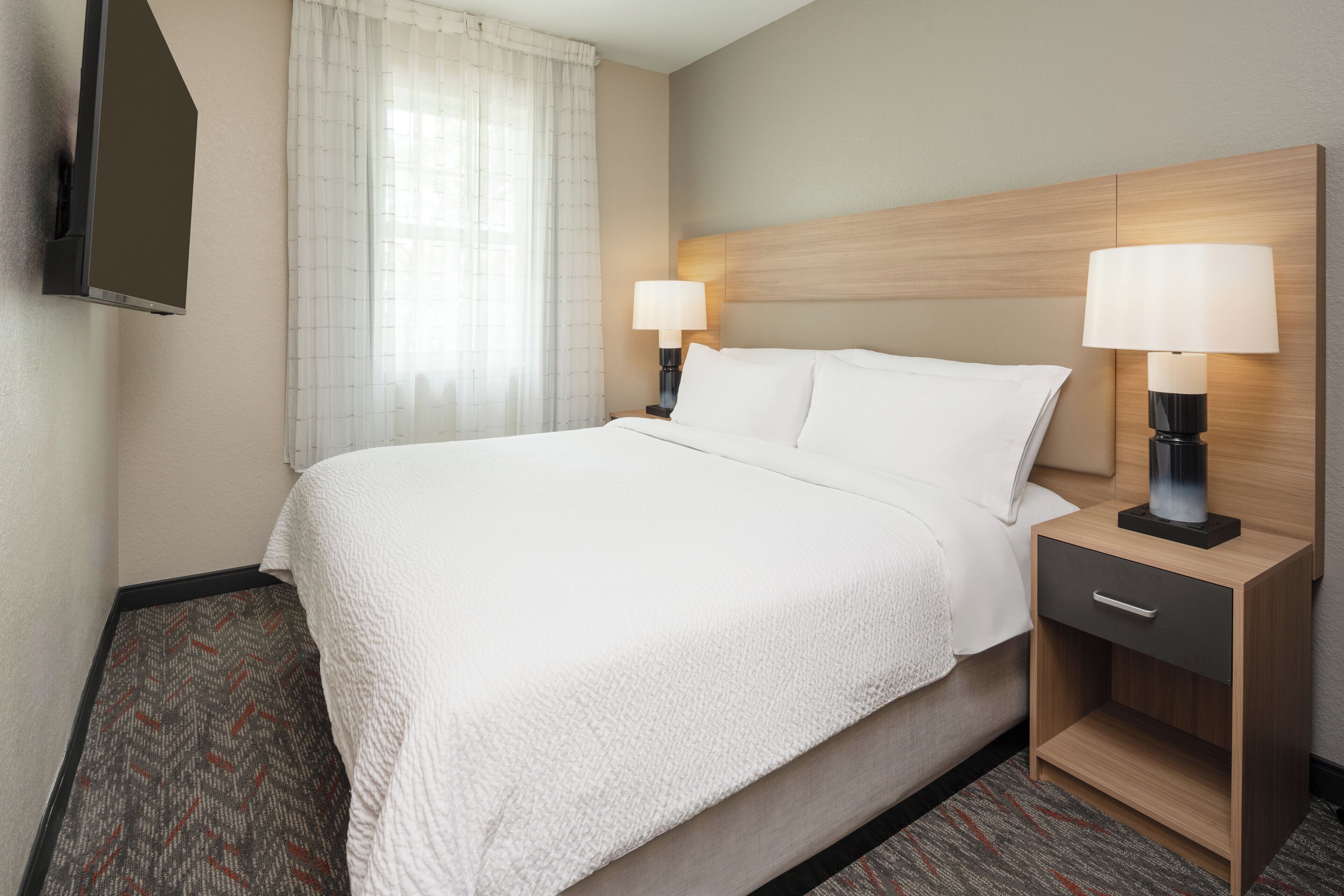 Une chambre d’hôtel avec un grand lit, deux tables de chevet avec des lampes, un téléviseur à écran plat fixé au mur et une fenêtre avec des rideaux légers.