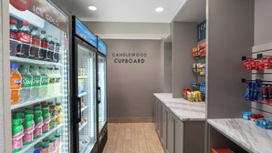 Käytävä, jossa on kylmälaitteella varustettu juomahylly, seinä, jossa on kyltti, jossa lukee ”CANDLEWOOD CUPBOARD”, sekä hyllyt, jotka on täytetty välipaloilla ja hygieniatuotteilla.