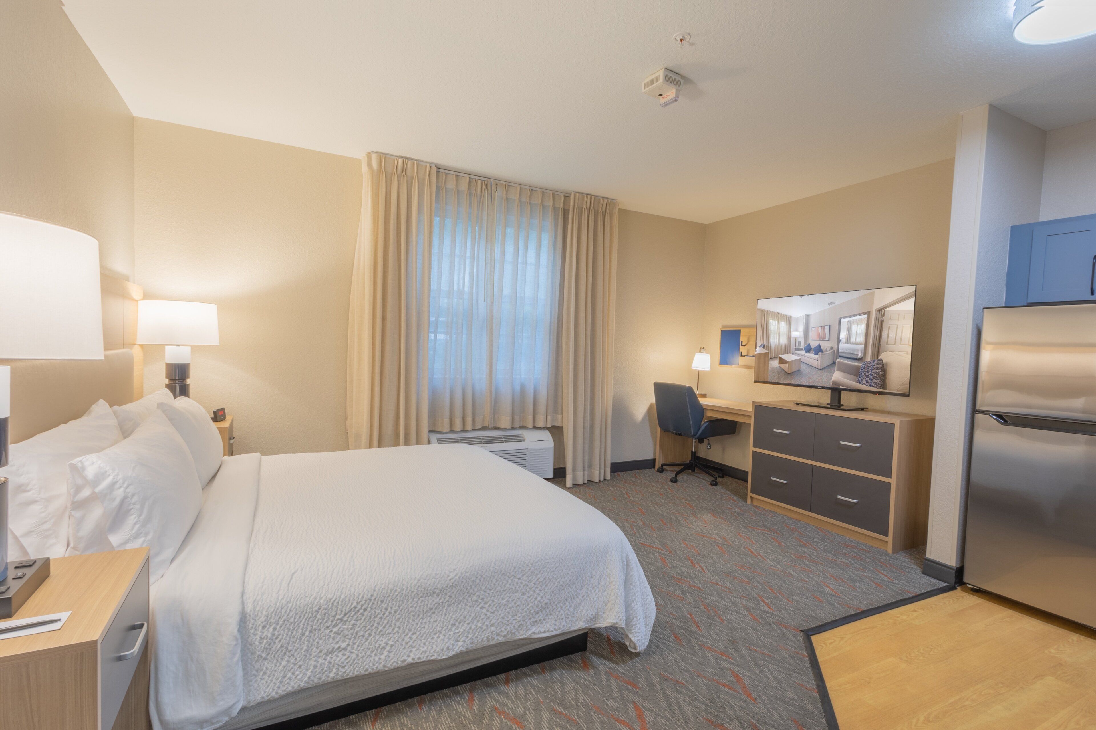 A hotel room with a bed, desk, chair, and refrigerator.