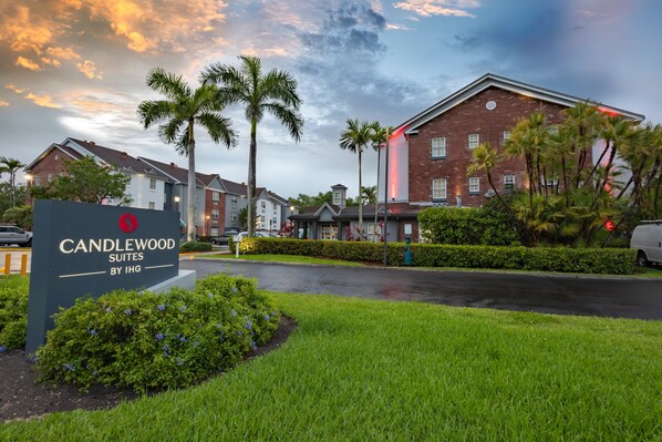 Entrada de un hotel con el letrero de Candlewood Suites by IHG, rodeada de follaje y palmeras.