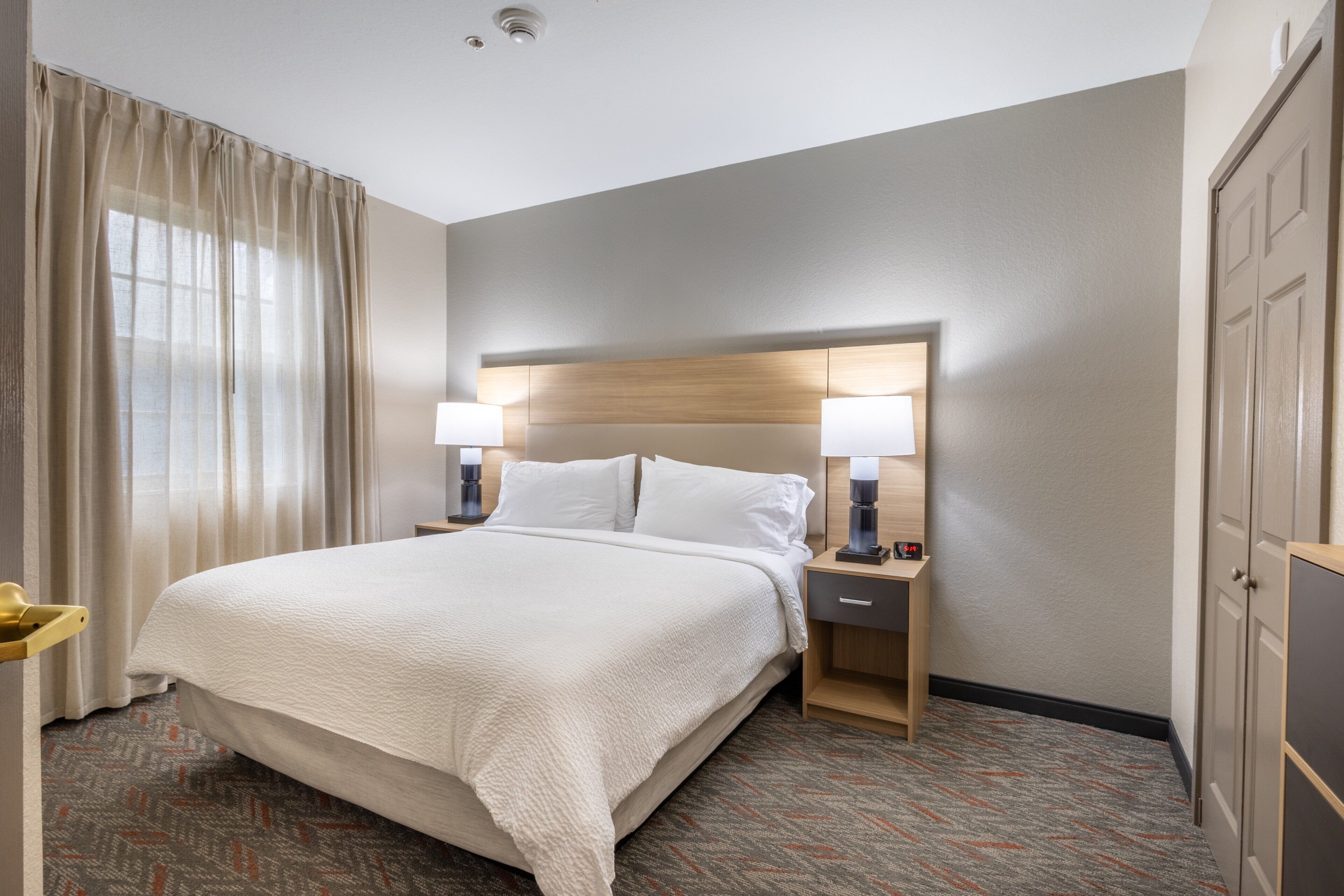 Una habitación de hotel con una cama grande, dos mesitas de noche con lámparas, un cabecero de madera, un armario y una alfombra estampada.