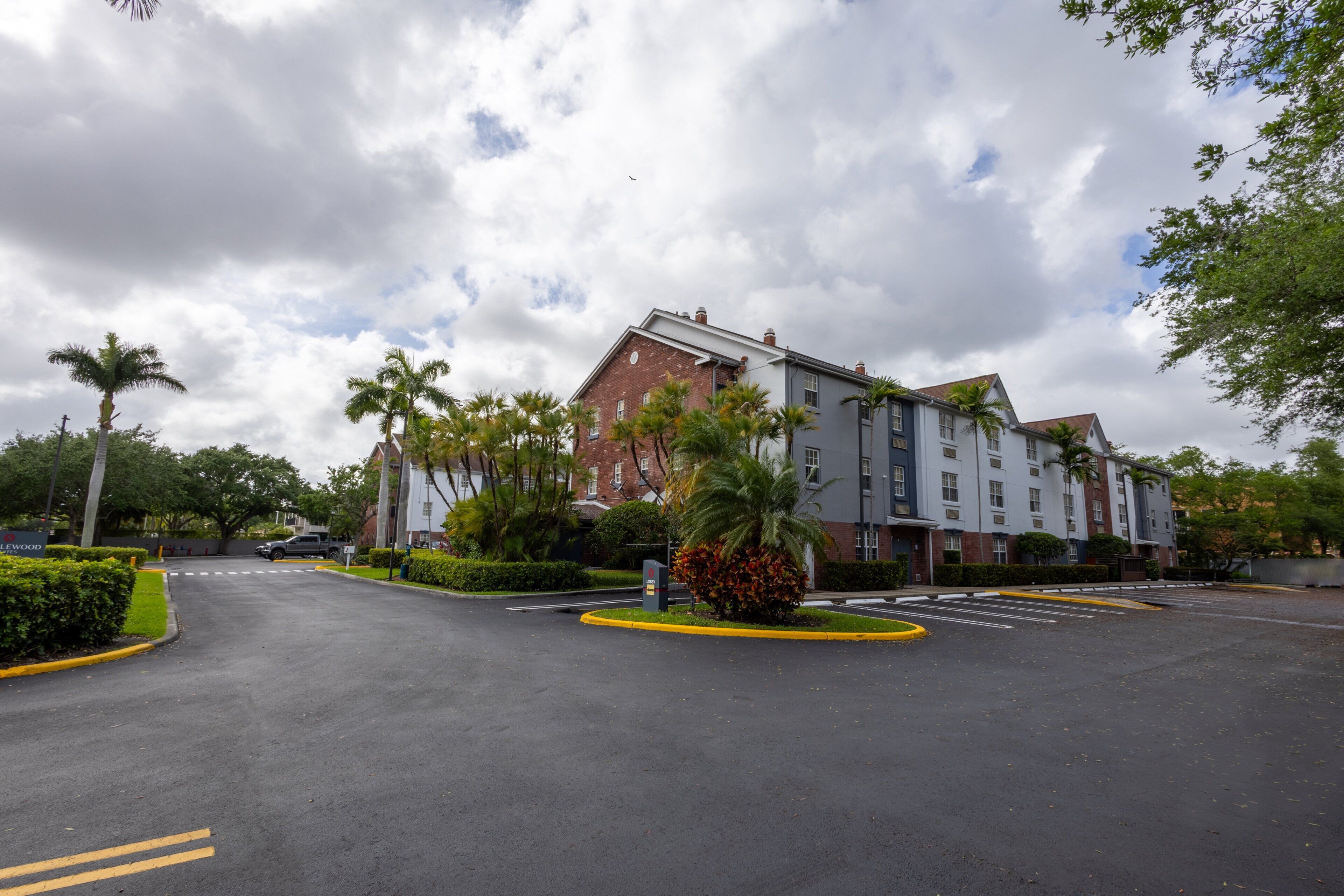 A multi-story building with a parking lot, palm trees, and a cloudy sky.